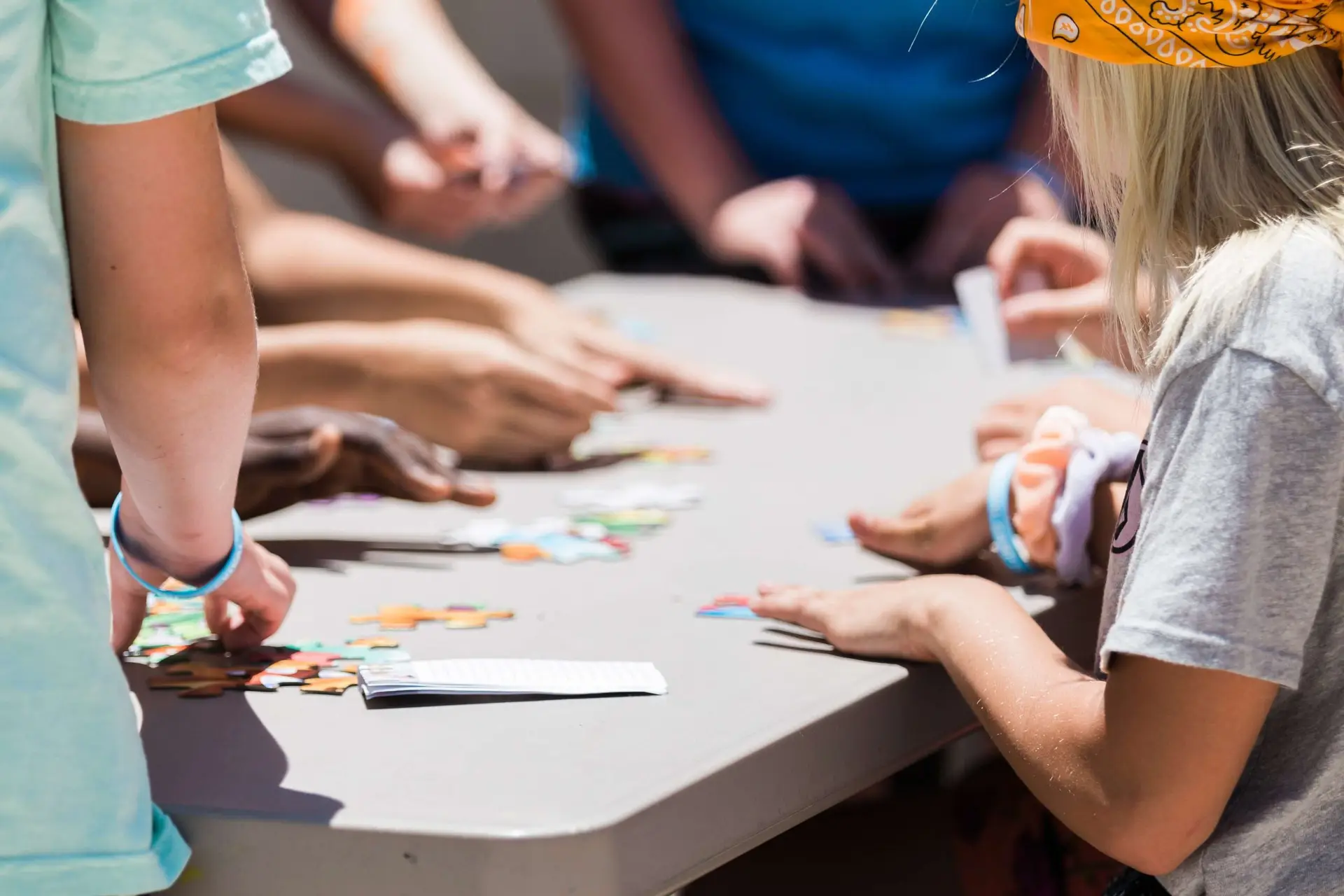 People gathered around a table outdoors, working on a jigsaw puzzle. Sunlight casts shadows on the group as they focus on assembling colorful pieces. A child with a yellow bandana enthusiastically shares sports trivia for kids while helping connect the pieces together.