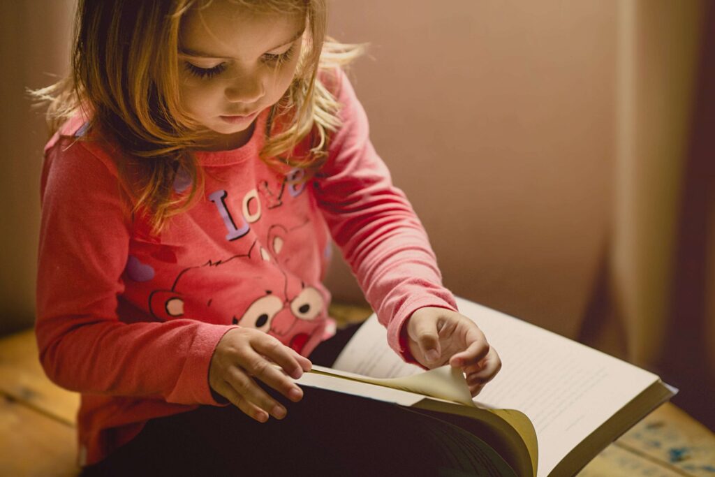 A young girl with long hair and a pink shirt, featuring cartoon bears and the word Love, sits on a wooden floor. She's engrossed in flipping through the pages of a mystery book for kids. The lighting is warm and soft, adding to the intrigue of her read.