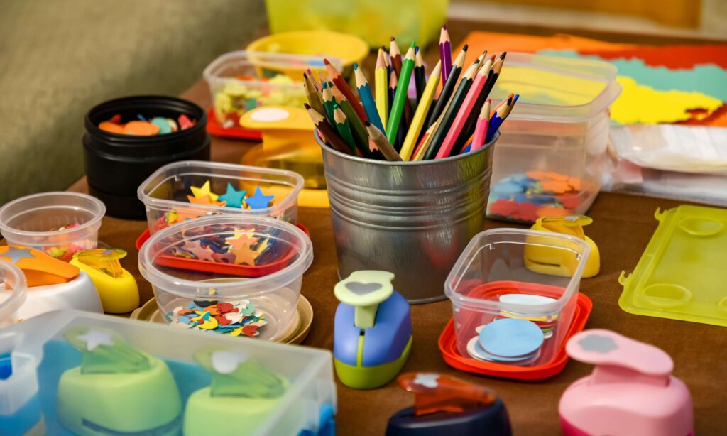 A table filled with arts and crafts supplies, including colorful pencils in a metal tin, various shaped paper punches, and containers of cut-out paper shapes. The vibrant items are perfect for creating friendship crafts, making it a lively and creative workspace.