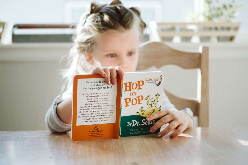 A young child with braided hair sits at a wooden table, reading Hop on Pop by Dr. Seuss. Sunlight filters through the window, illuminating the educational book's colorful cover and the child's focused expression.