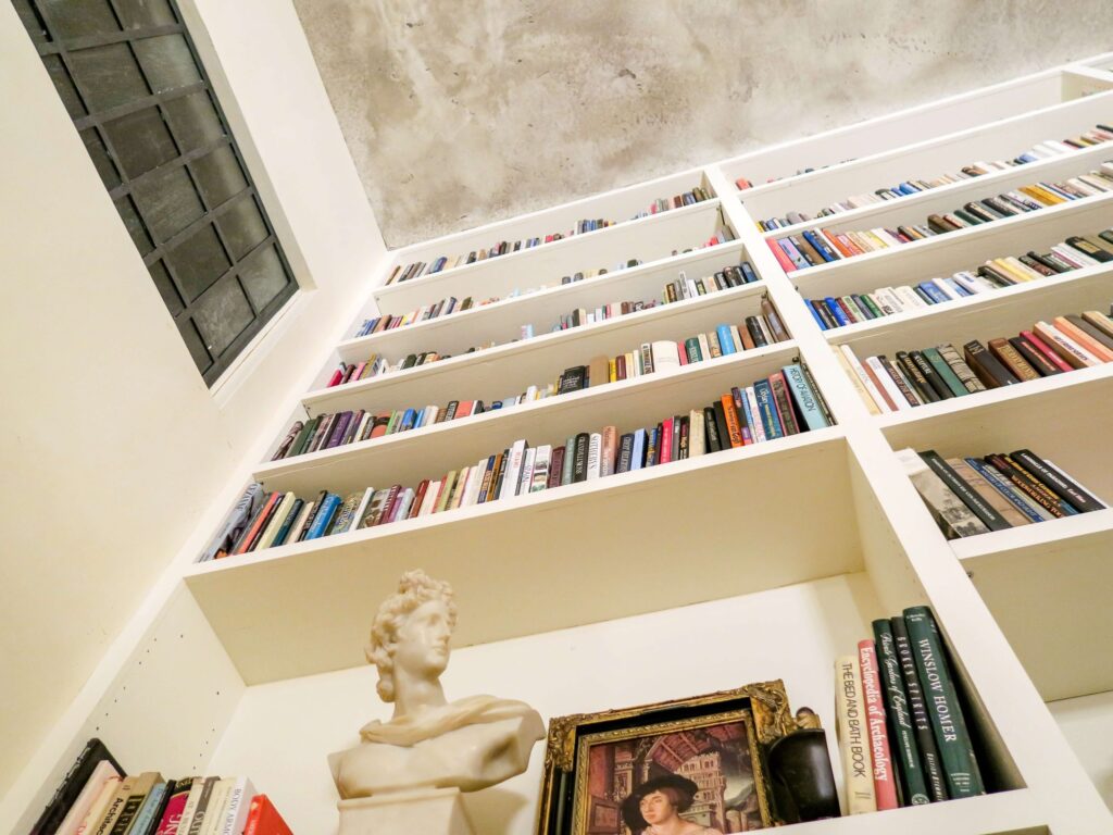 Low-angle view of a home library with tall white bookshelves filled with books, including history books for kids. A bust sculpture and a framed portrait are displayed on a lower shelf. The ceiling features a textured concrete finish, and a window graces the left wall.