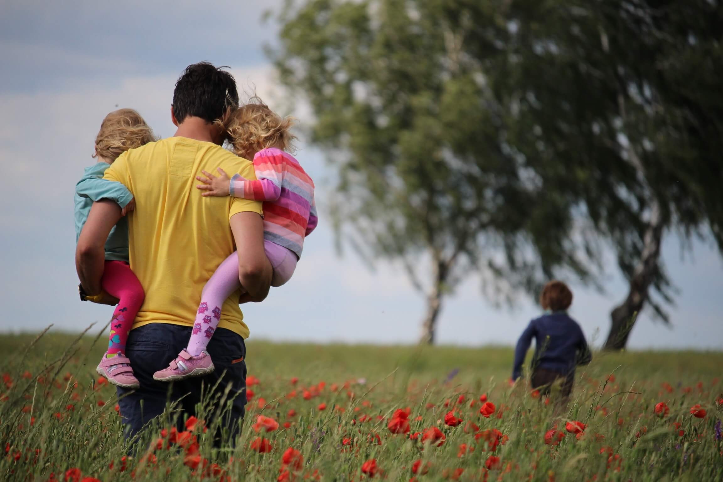 A man in a yellow shirt carries two young children through a field of red poppies, reminiscent of scenes from beloved Christian TV shows for kids. Another child runs ahead into the green grass under a cloudy sky, as trees sway in the breeze in the background.