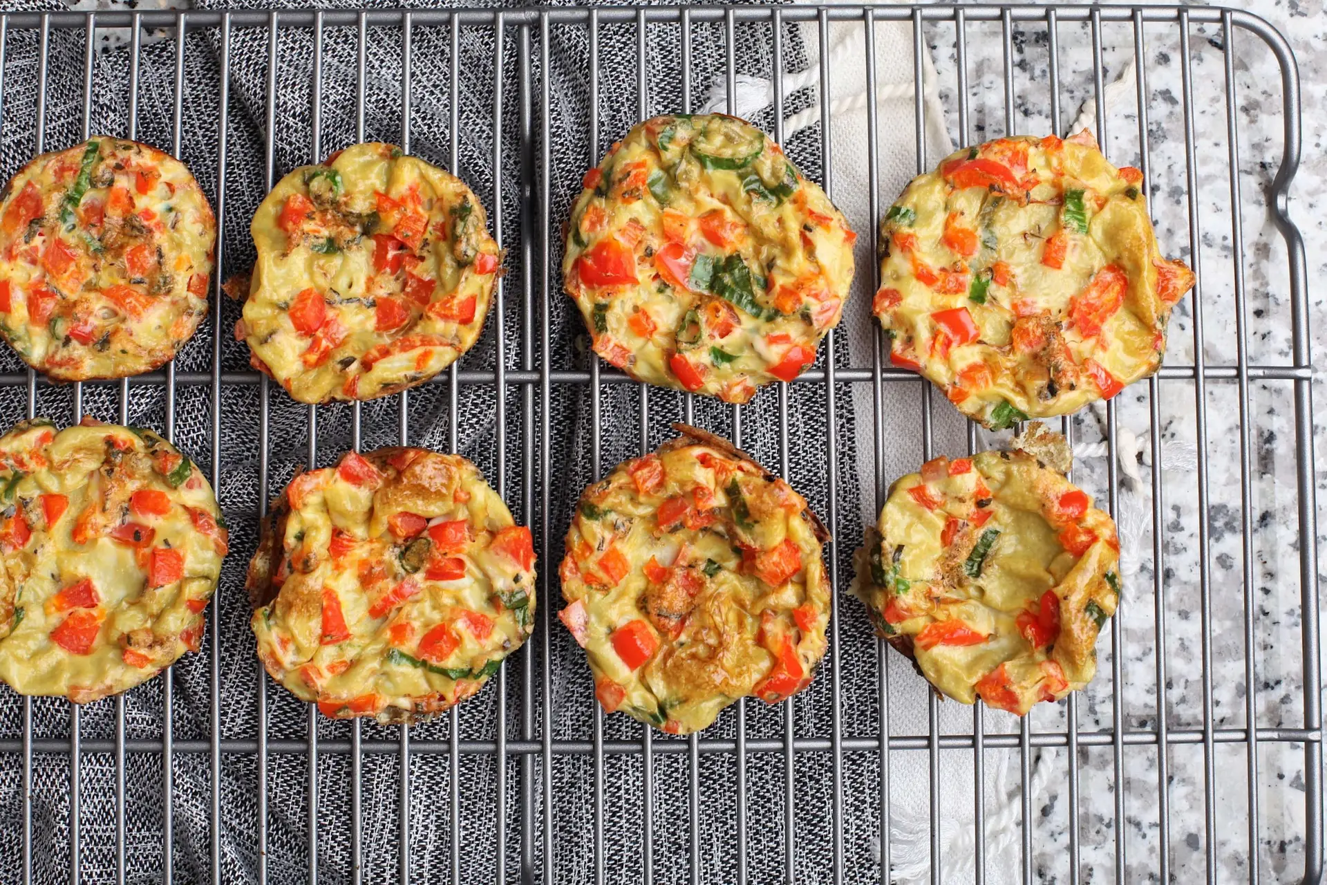 Eight mini vegetable omelets with diced red peppers, zucchini, and onions are cooling on a wire rack over a textured surface. These golden brown delights, perfect for toddler breakfast ideas, are arranged neatly in two rows.