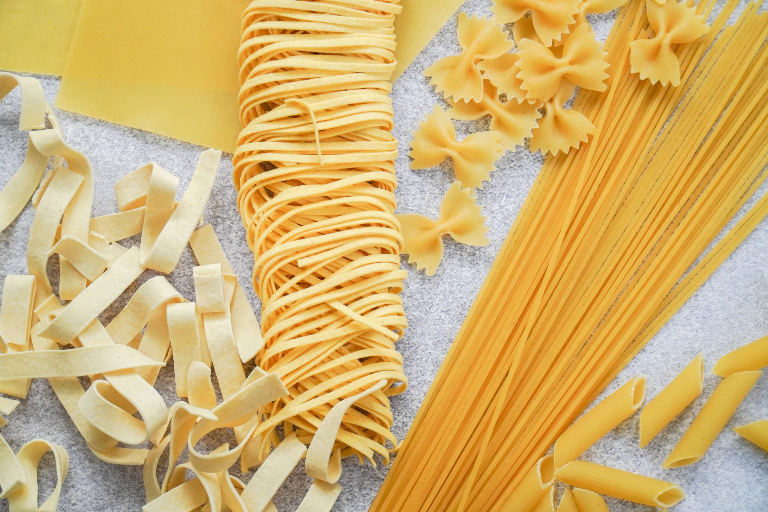 An assortment of uncooked pasta types on a textured surface, including fettuccine, spaghetti, lasagna sheets, farfalle, and tagliatelle. These kid-friendly pasta favorites showcase a variety of shapes and sizes in shades of light yellow.