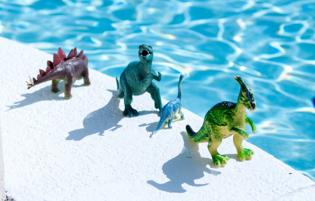 Four toy dinosaurs in different colors stand on the edge of a bright blue swimming pool, perfect dinosaur gifts for kids. The dinosaurs cast distinct shadows on the white poolside surface.