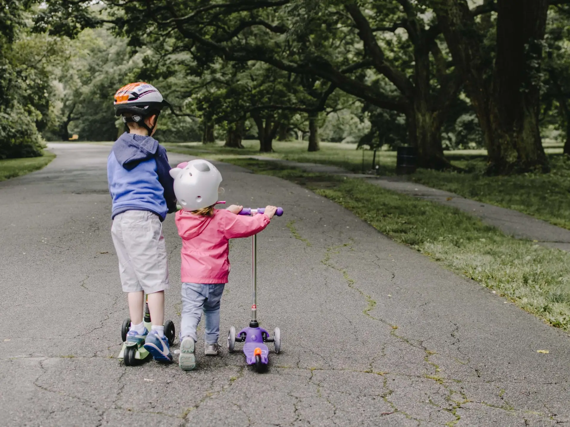 Two children wearing helmets ride scooters, the perfect summer toys for kids, down a paved path in a park. The older child sports a blue jacket and gray shorts, while the younger wears a pink jacket and blue pants. They are surrounded by lush green trees.