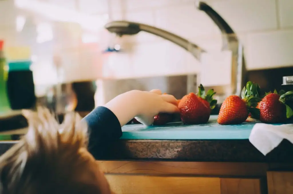 A child reaches for gluten-free snacks on a kitchen counter next to a sink. The strawberries sit on a blue cutting board, with their hand eagerly extended toward them. The background features a softly blurred kitchen setting.