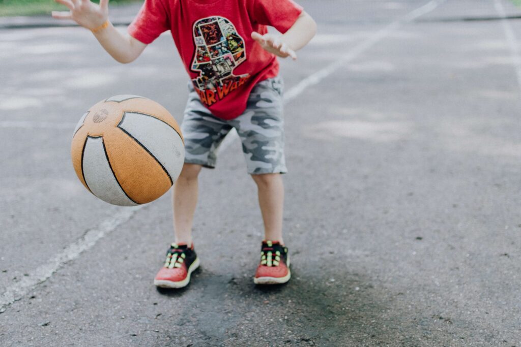 A child in a red T-shirt and camouflage shorts enjoys playing basketball outdoors, one of the best sports for kids. The ball bounces energetically on the paved surface, though the child’s face remains unseen.