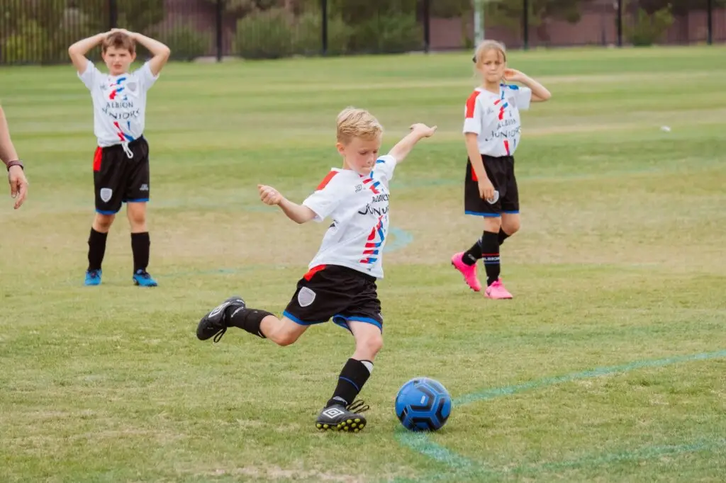 A young boy in a white soccer uniform is kicking a blue soccer ball on a grassy field, showcasing why kids should play sports. Two other children, in similar uniforms, stand in the background watching. They are all part of a youth soccer team.