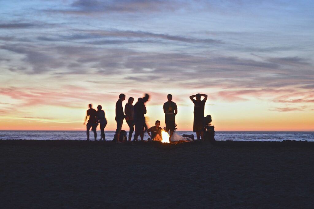 A group of people are gathered around a campfire on a beach at sunset, enjoying lively campfire games. The sky is painted with hues of orange, pink, and blue, and the silhouettes of the group are visible against the twilight backdrop.