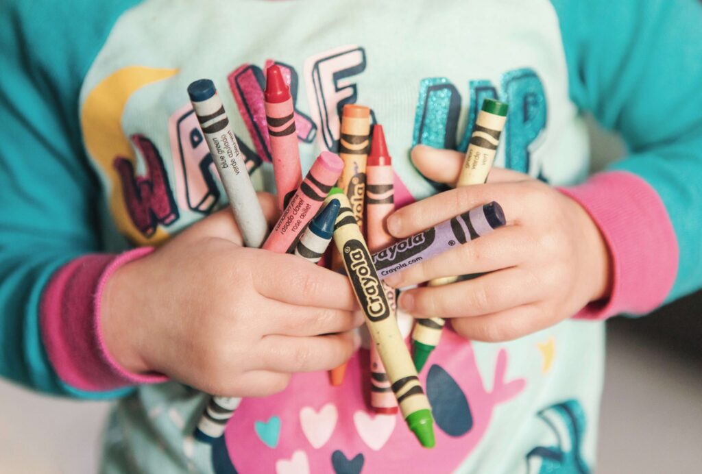 A child in a colorful shirt holds a bunch of Crayola crayons, featuring various colors like red, green, and blue. With playful designs and lettering on the shirt, it creates a cheerful and artistic atmosphere—perfect for inspiring free indoor activities for kids.