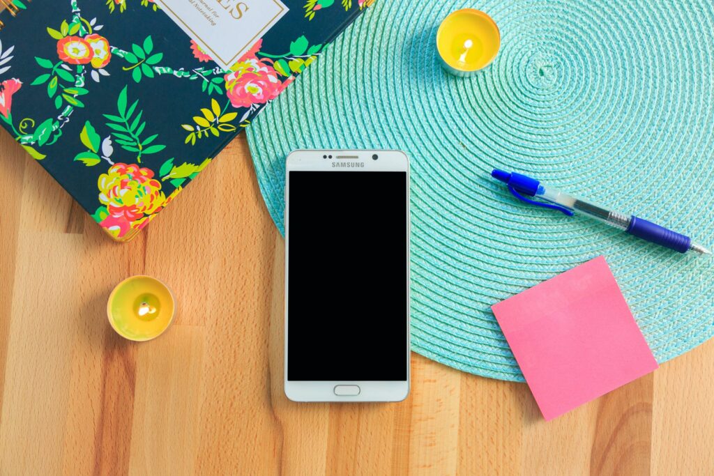 A smartphone with a black screen rests on a turquoise woven mat on a wooden table, embodying the mindful theme of Internet Safety Month. Nearby are a colorful floral notebook, a blue pen, a pink sticky note, and two small lit candles in yellow holders.