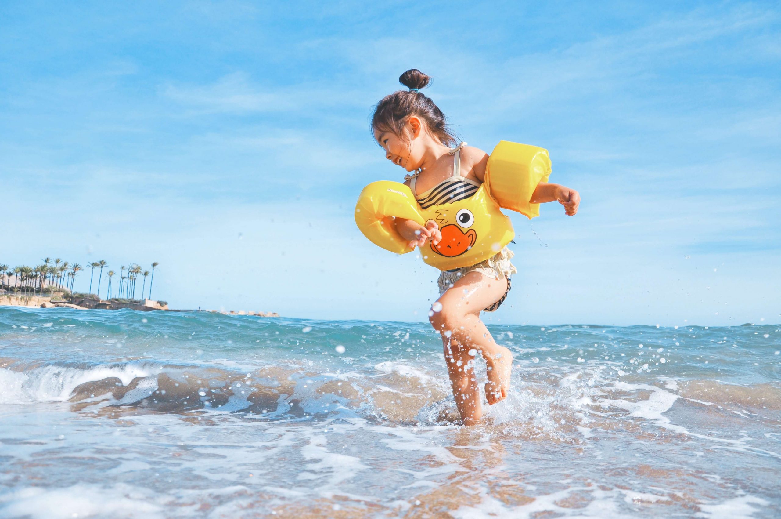 A young girl, embodying the essence of outdoor toddler activities, wears yellow arm floaties as she plays joyfully at the beach, splashing in the waves. The sky is clear and blue, with palm trees swaying gently in the background.