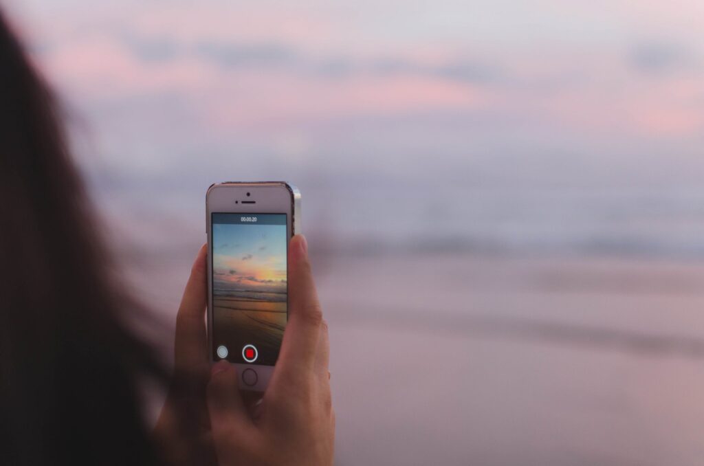 As part of National Cell Phone Courtesy Month, a person holds a smartphone, discreetly recording the serene beach sunset. The screen reveals soft orange and pink hues mirrored on calm waters, crafting a peaceful and picturesque atmosphere for all to enjoy without disturbance.