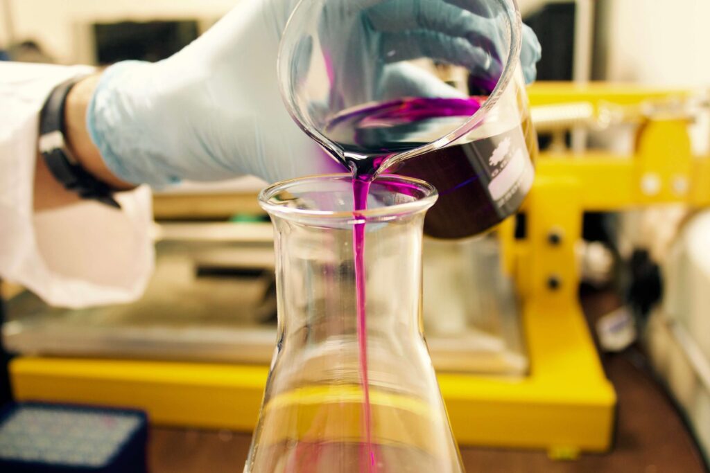 A person in a lab coat and gloves carefully pours a bright purple liquid from a beaker into a conical flask, reminiscent of scenes from science movies for kids. In the background, an array of laboratory equipment is neatly arranged on the workbench.