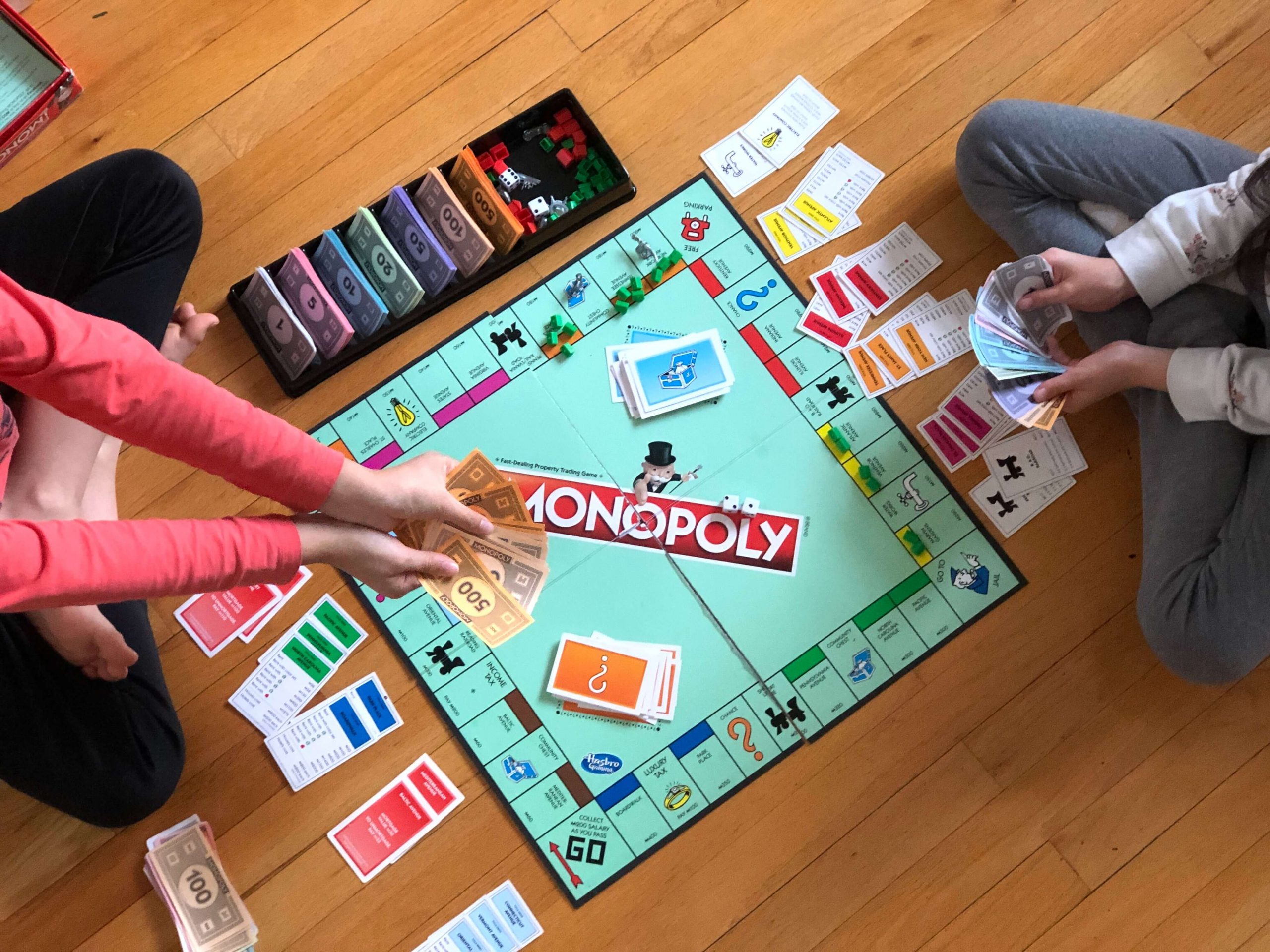 Two people playing Monopoly on a wooden floor. The game board is set up, with cards, houses, hotels, and money scattered around. Embracing the spirit of educational board games for kids, one person holds a stack of money while the other organizes property cards.