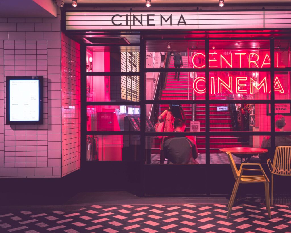 Brightly lit cinema entrance with a neon Central Cinema sign, glass doors, and red interior lighting. A person stands at the counter discussing new kids movies of 2023. A small round table with chairs is visible in the foreground on a tiled floor.