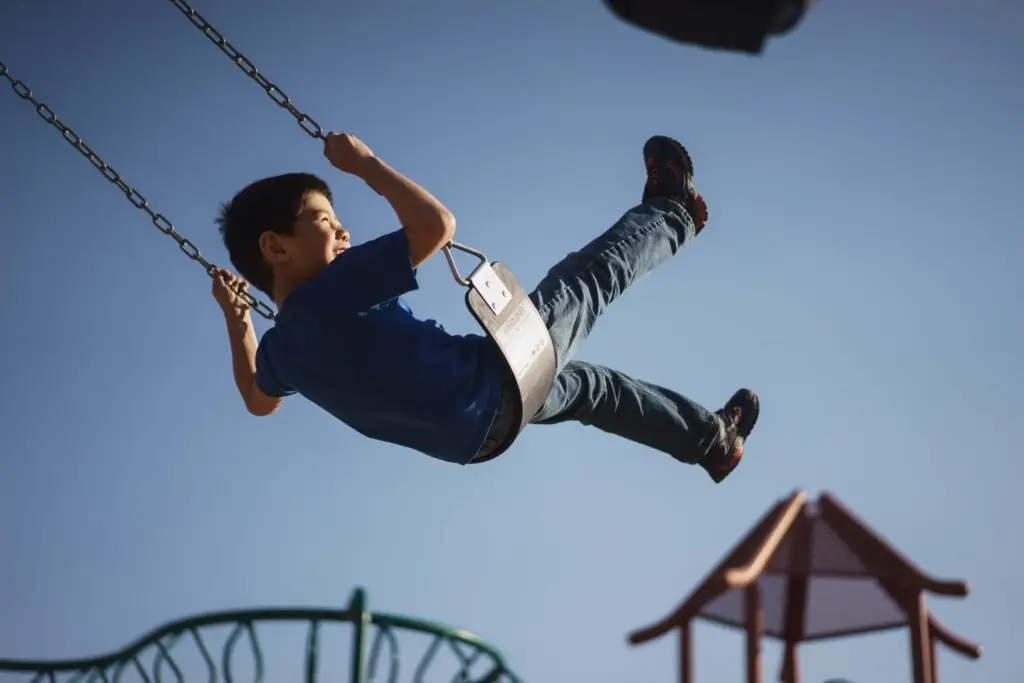 A young boy joyfully swings high in the air at a playground, embodying the spirit of books about self-confidence. Wearing a blue shirt and jeans, he is captured mid-swing against a clear blue sky. In the background, playground equipment is visible.