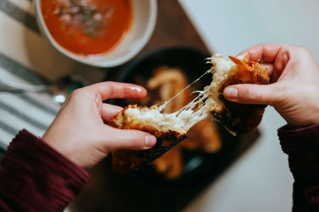 A person pulls apart a grilled cheese sandwich, revealing melted cheese strands. A bowl of tomato soup is in the background, alongside a black plate of food. Perfect for kid-friendly air fryer recipes, the striped tablecloth adds a cozy touch to this delicious scene.