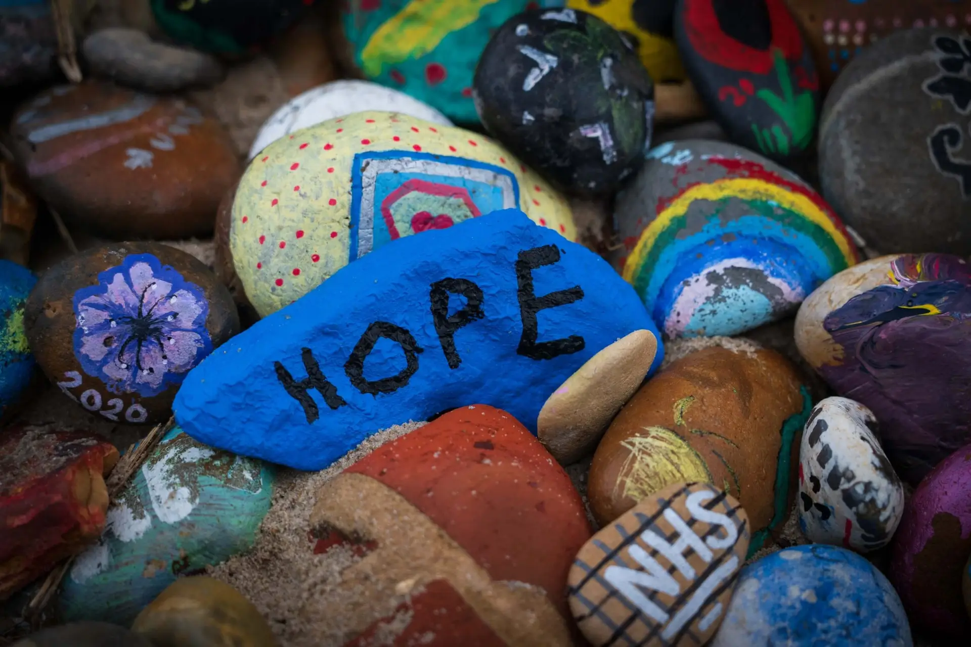 A collection of painted stones, featuring a prominent blue one with HOPE written on it. Perfect for kindness activities for kids, other stones showcase colorful designs like rainbows, flowers, and NHS painted in white, spreading positivity and creativity.