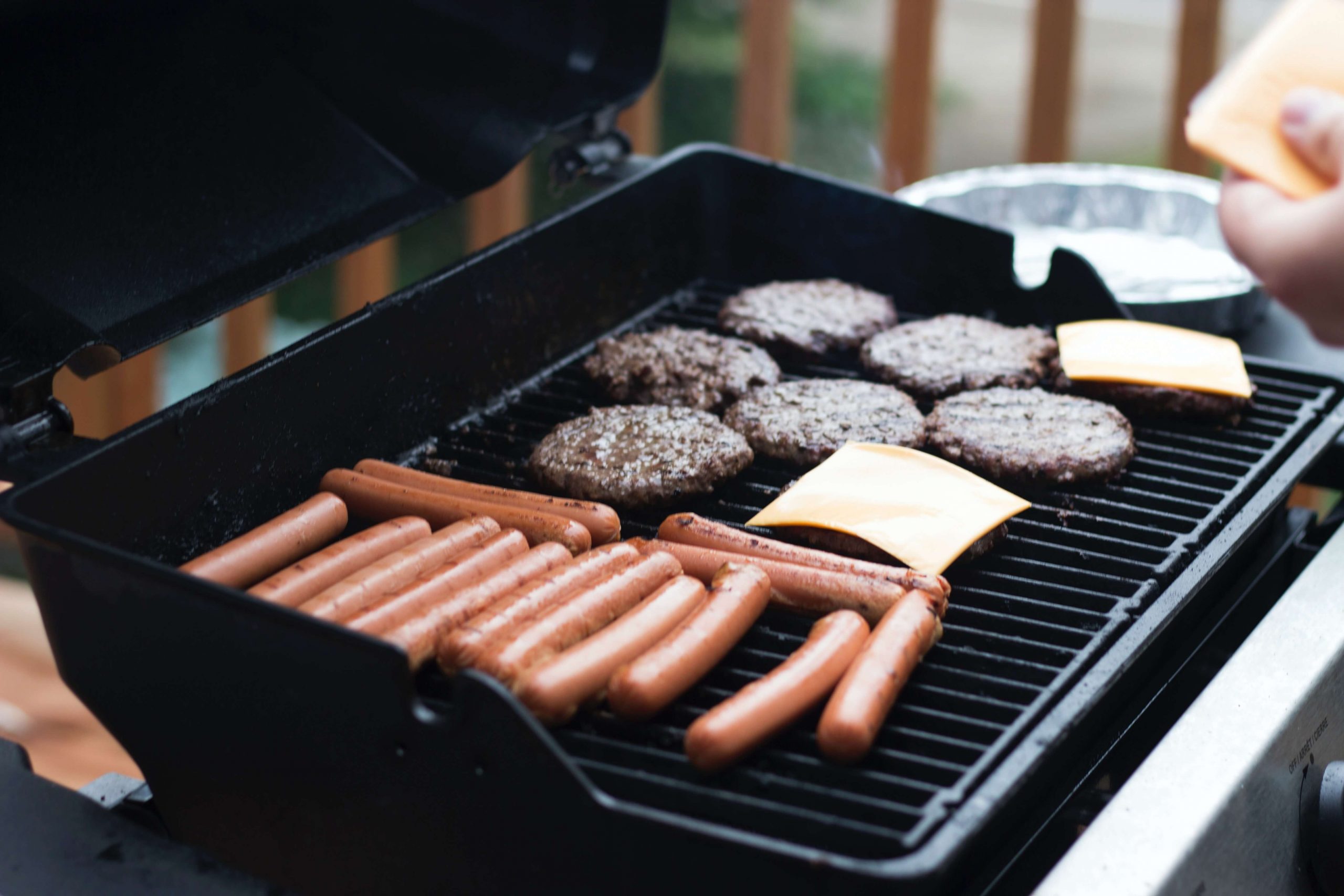 A grill loaded with hot dogs and hamburger patties, some with cheese melting on top, evokes the cozy vibe of campfire-themed party food. The lid is open, revealing the sizzling feast and suggesting a rustic backyard barbecue setting.