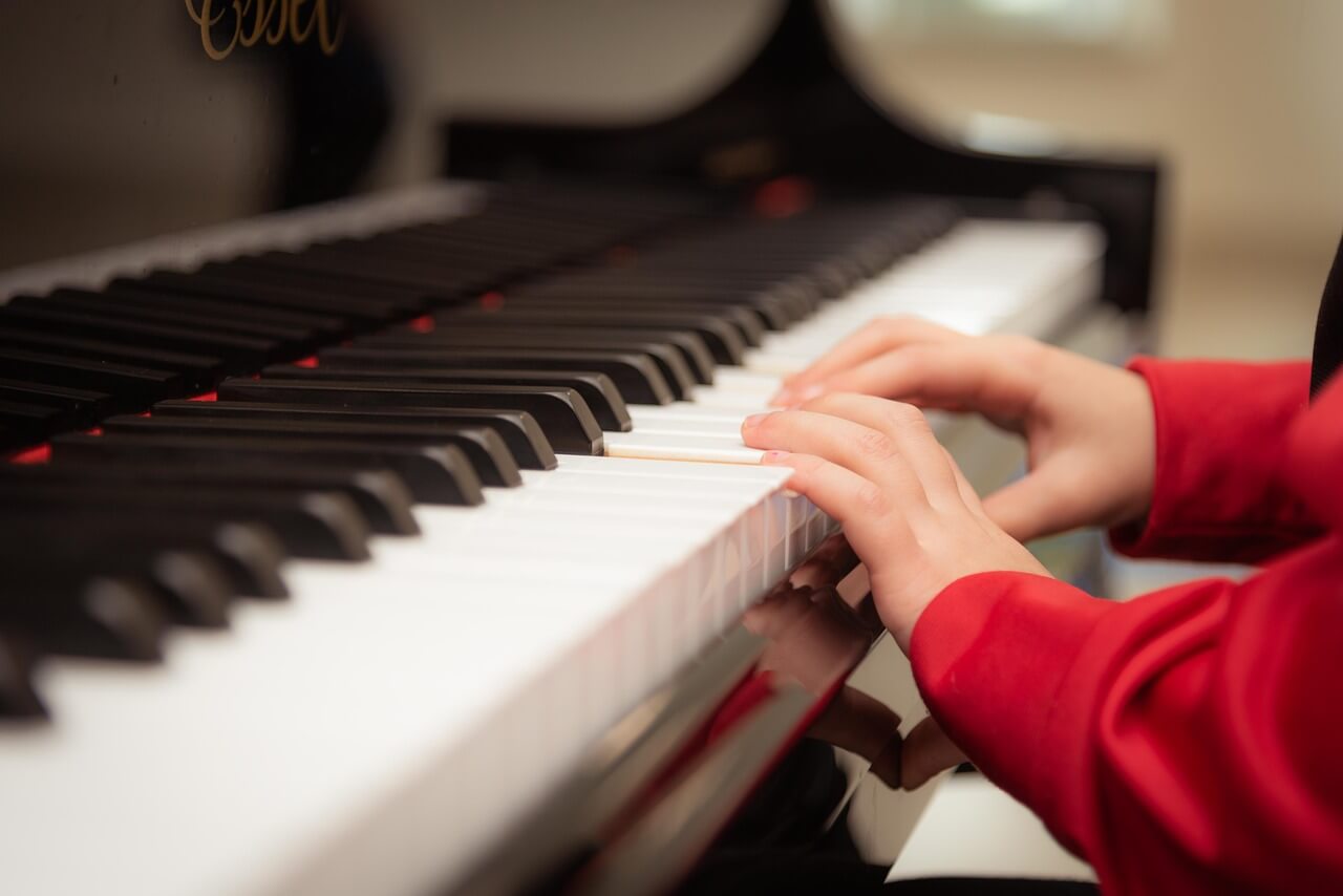 Close-up of a person in a red shirt playing a black and white piano, reminiscent of engaging piano games for kids. The hands are positioned on the keys, suggesting they are in mid-performance. The background is slightly blurred, emphasizing the focus on the hands and keys.