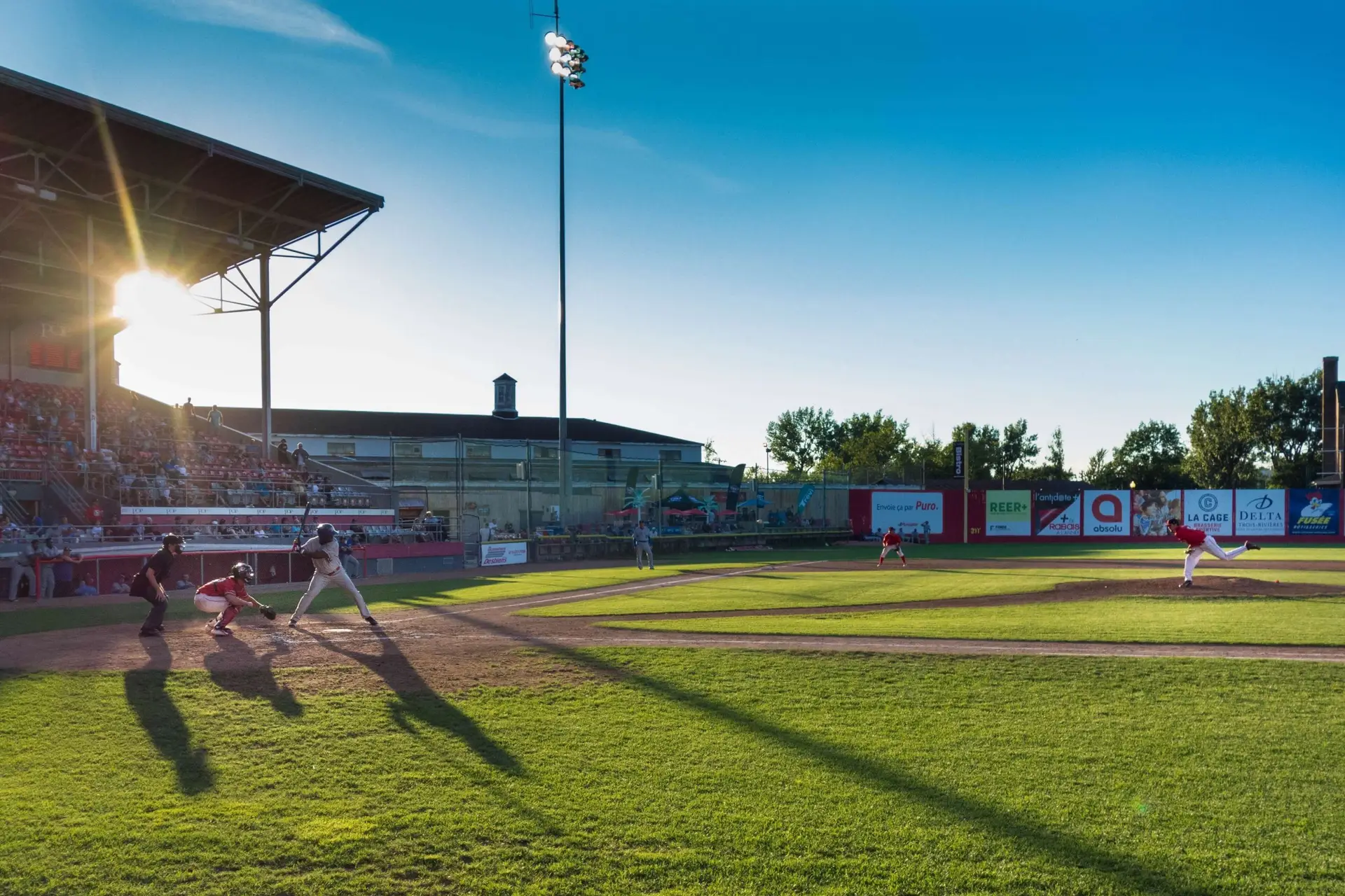 As the sun sets on a thrilling baseball game, reminiscent of scenes from the best kids sports movies, a batter, catcher, and umpire hold their ground. Spectators fill the stadium, while long shadows stretch across the vibrant green field under bright lights against a clear sky.