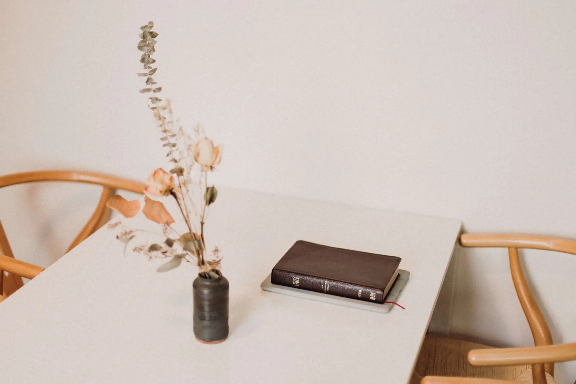 A minimalistic table setup with a closed book on the right and a small black vase with dried flowers on the left. Two wooden chairs, perfect for family discussions about Christian movies for kids, are partially visible. The backdrop is a plain white wall.