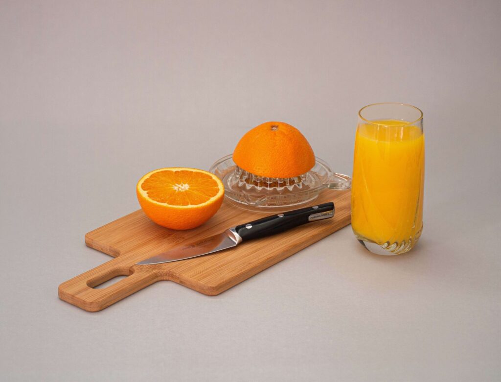 A halved orange rests on a wooden cutting board alongside a knife and citrus juicer, ready to create one of the perfect summer drinks for kids. A glass of freshly squeezed orange juice stands invitingly nearby against the neutral gray background.