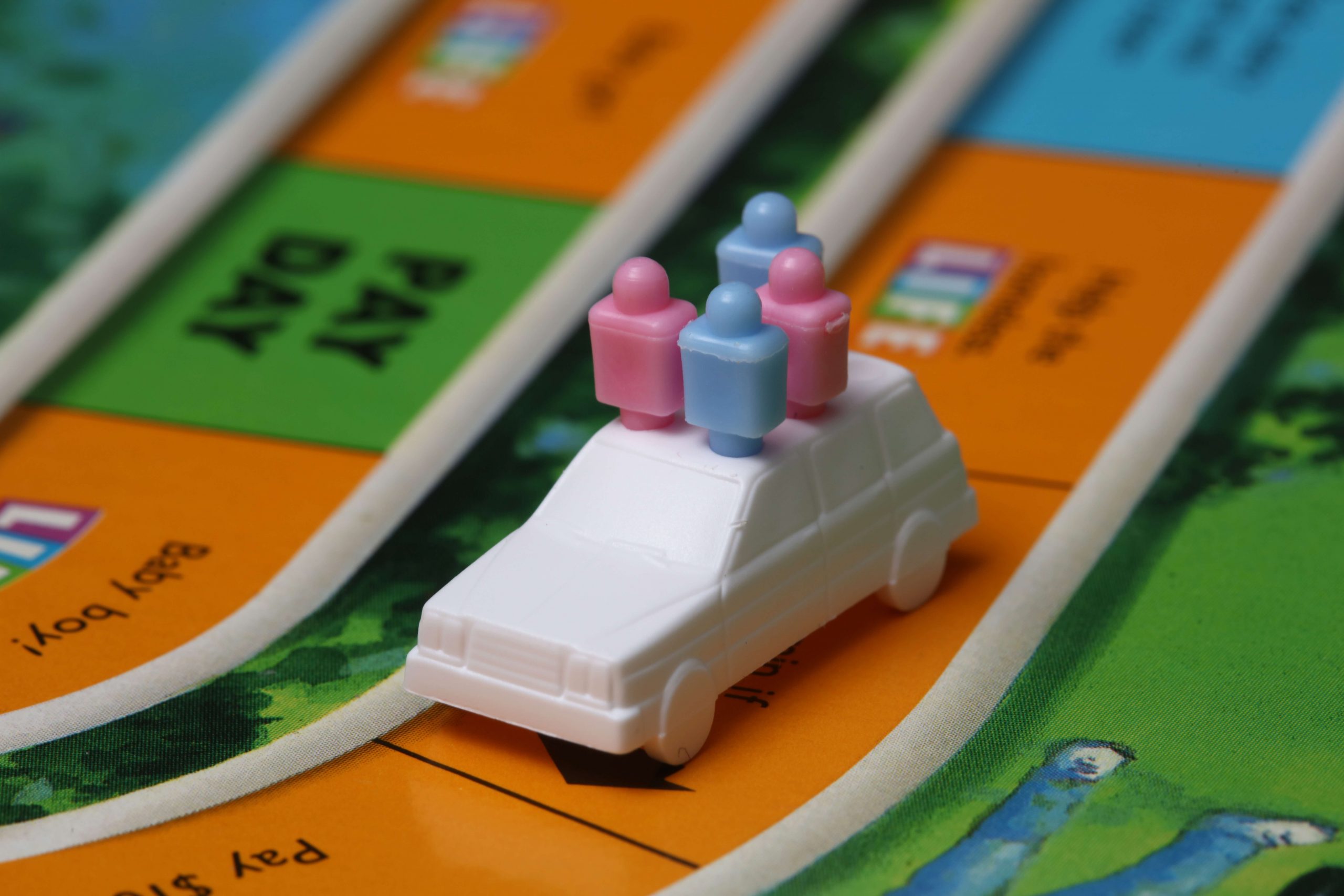 Close-up of a plastic game piece, shaped like a white car, on a board game. This brain game for kids features the car with four peg-shaped passengers, two blue and two pink, parked on an orange space labeled PAY DAY. Surrounding spaces are multicolored with text.