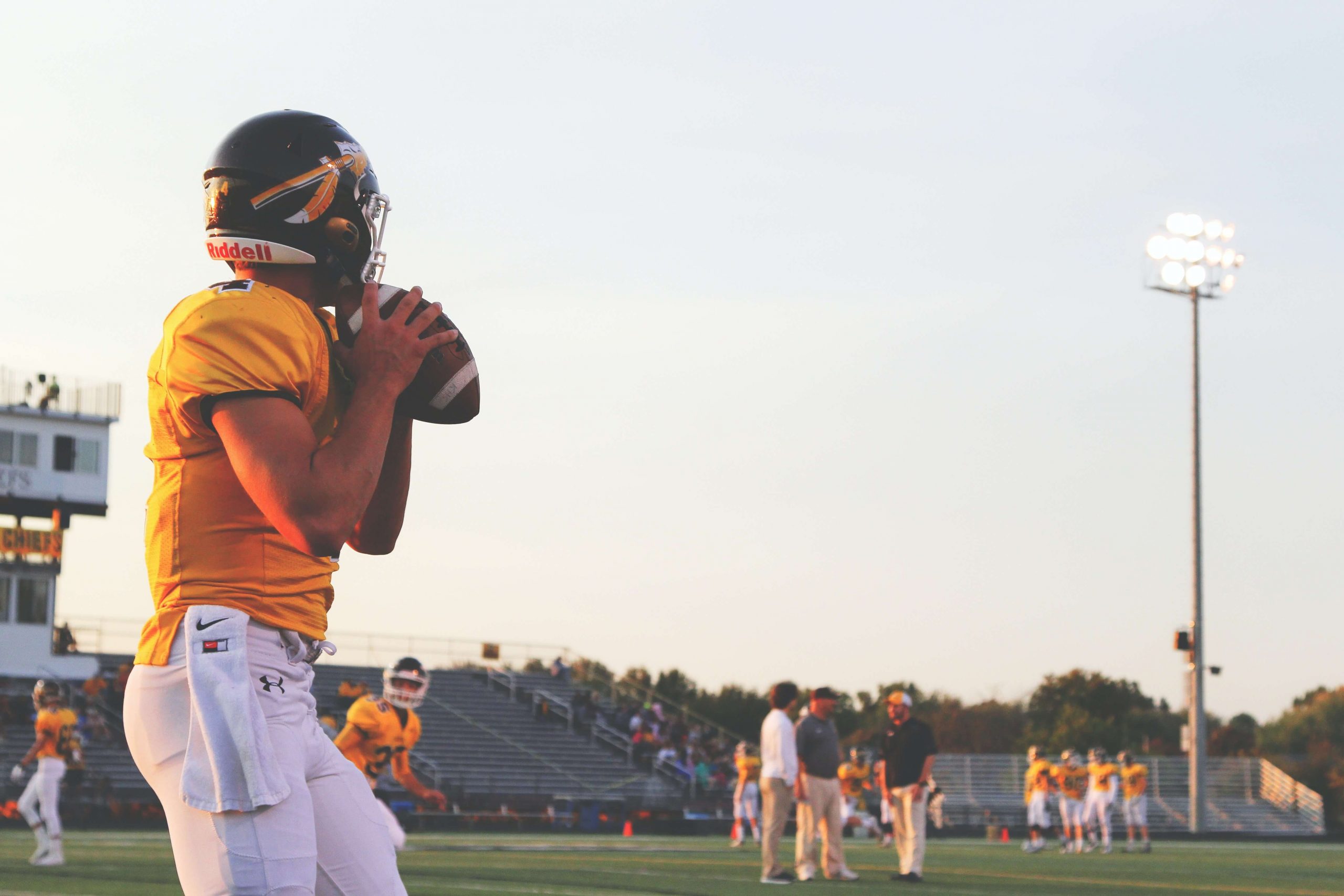 A football player in a yellow jersey holds a football, preparing to throw during a practice under the stadium lights. Other players and coaches can be seen in the background, creating a scene reminiscent of inspiring football movies for kids.