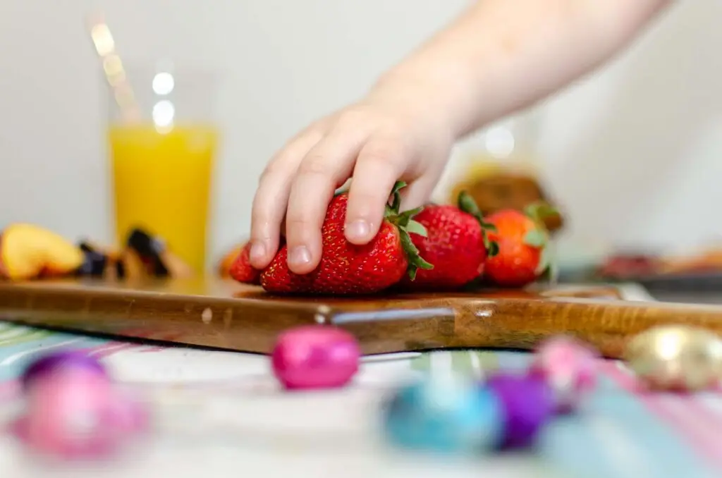 A child's hand reaches for strawberries on a wooden board. In the background, there's a glass of orange juice and dairy-free snacks, with colorful foil-wrapped chocolates scattered on a striped tablecloth.