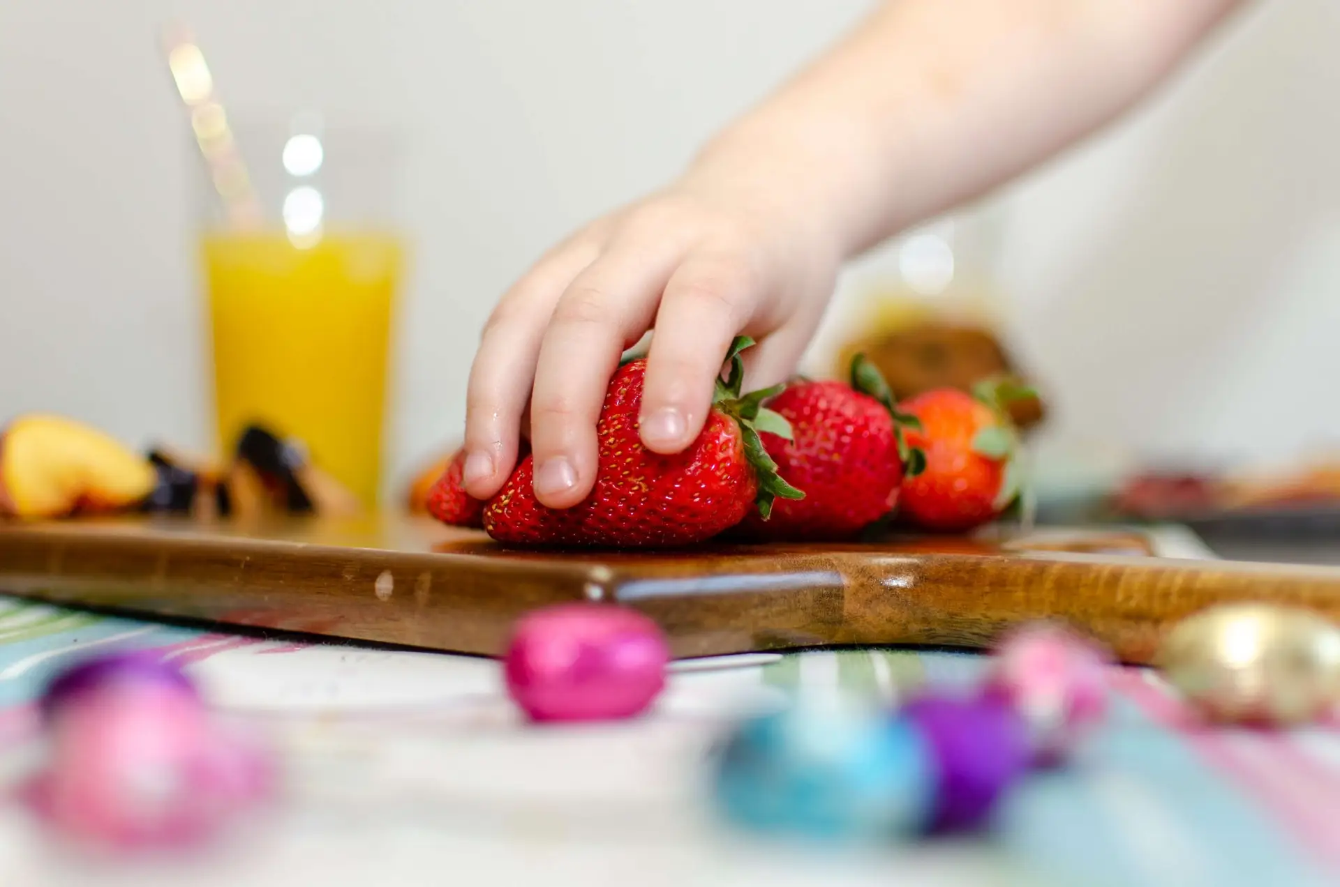 A child's hand reaches for strawberries on a wooden board. In the background, there's a glass of orange juice and dairy-free snacks, with colorful foil-wrapped chocolates scattered on a striped tablecloth.