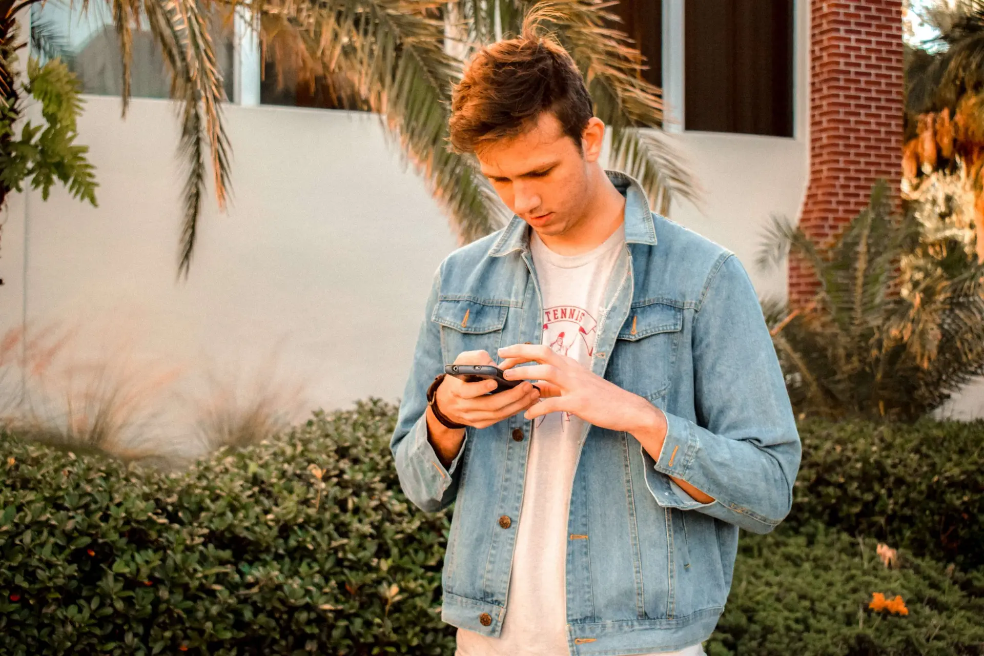 A person in a denim jacket checks their smartphone, contemplating the average screen time for teens, as they stand amidst lush greenery with palm trees and a building in the background. The sunlight casts a warm glow on the scene.