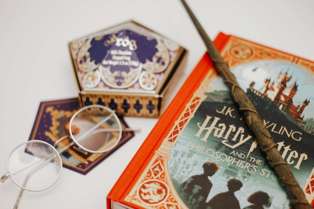 A perfect Harry Potter gift for kids, this charming setup features a copy of Harry Potter and the Philosopher's Stone with a wand resting on it, accompanied by iconic round glasses and a chocolate frog box.