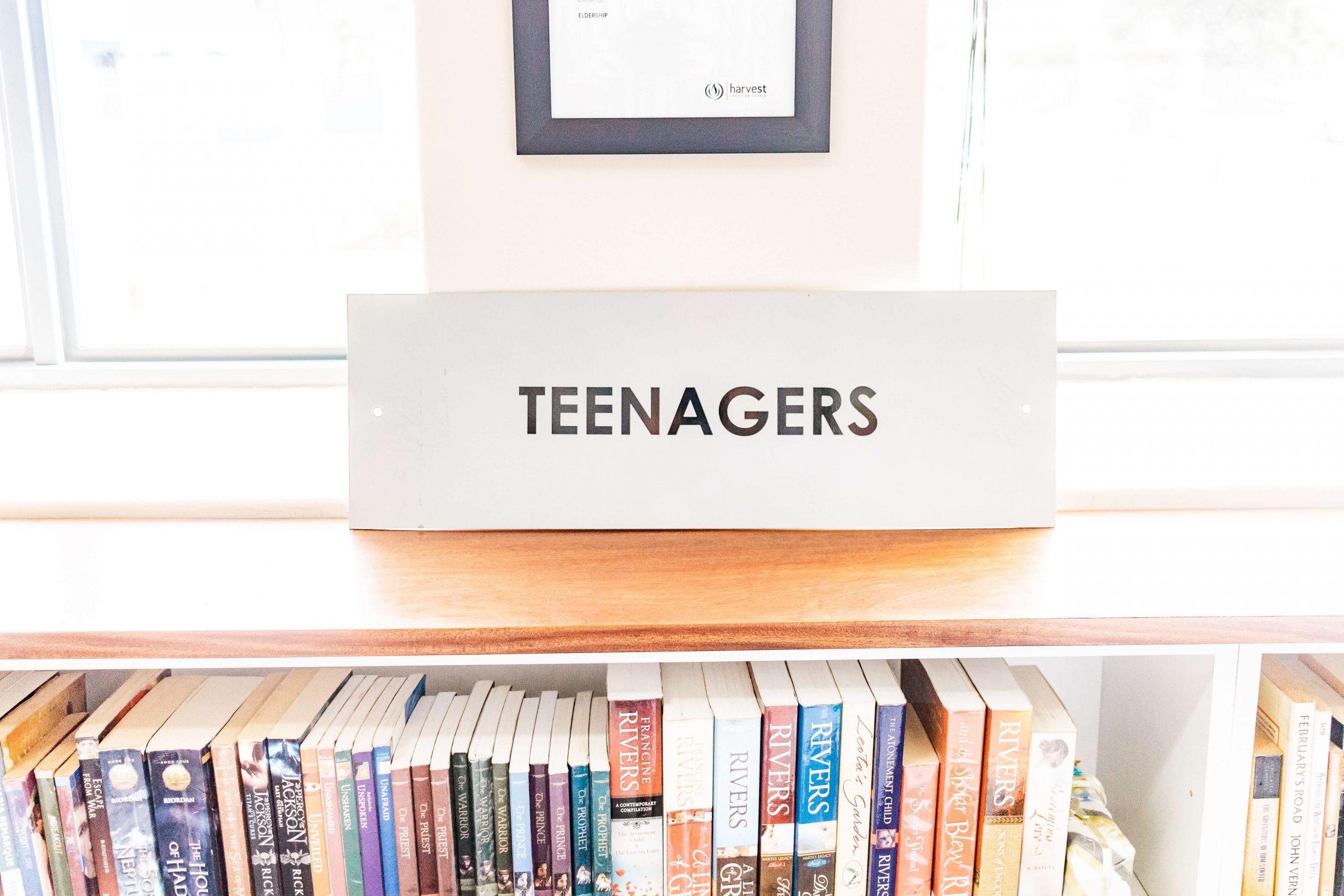 A shelf in a library labeled TEENAGERS holds a diverse array of books, mostly colorful paperbacks. Nearby, a framed certificate featuring a parenting contract subtly encourages positive dialogue.