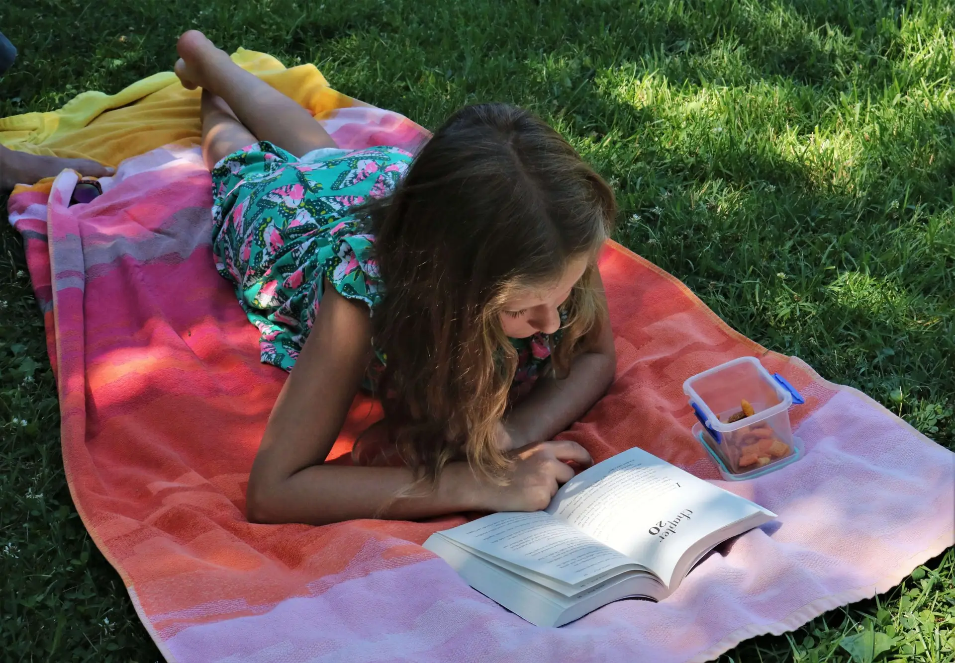 A young girl lies on her stomach on a colorful towel spread over the grass, immersed in a summer book for kids. She's wearing a green floral dress, and there's a small container with snacks beside her. Sunlight dapples through the trees, casting soft shadows.