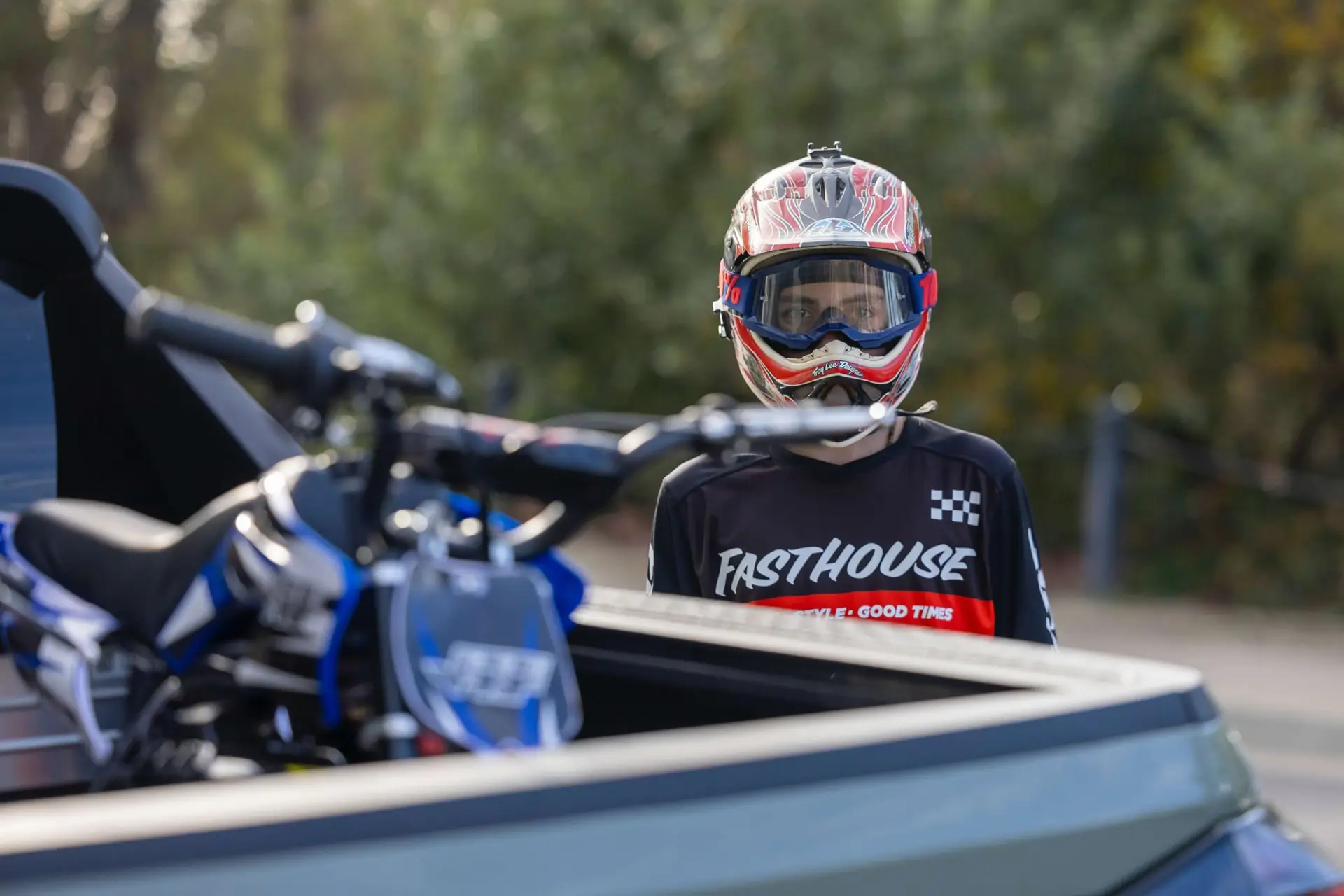 A person wearing a red and white helmet and a black Fasthouse jersey stands by a truck with a blue dirt bike in the back, perfect for inspiring dreams of kids' dirt bikes. The background features blurred trees.