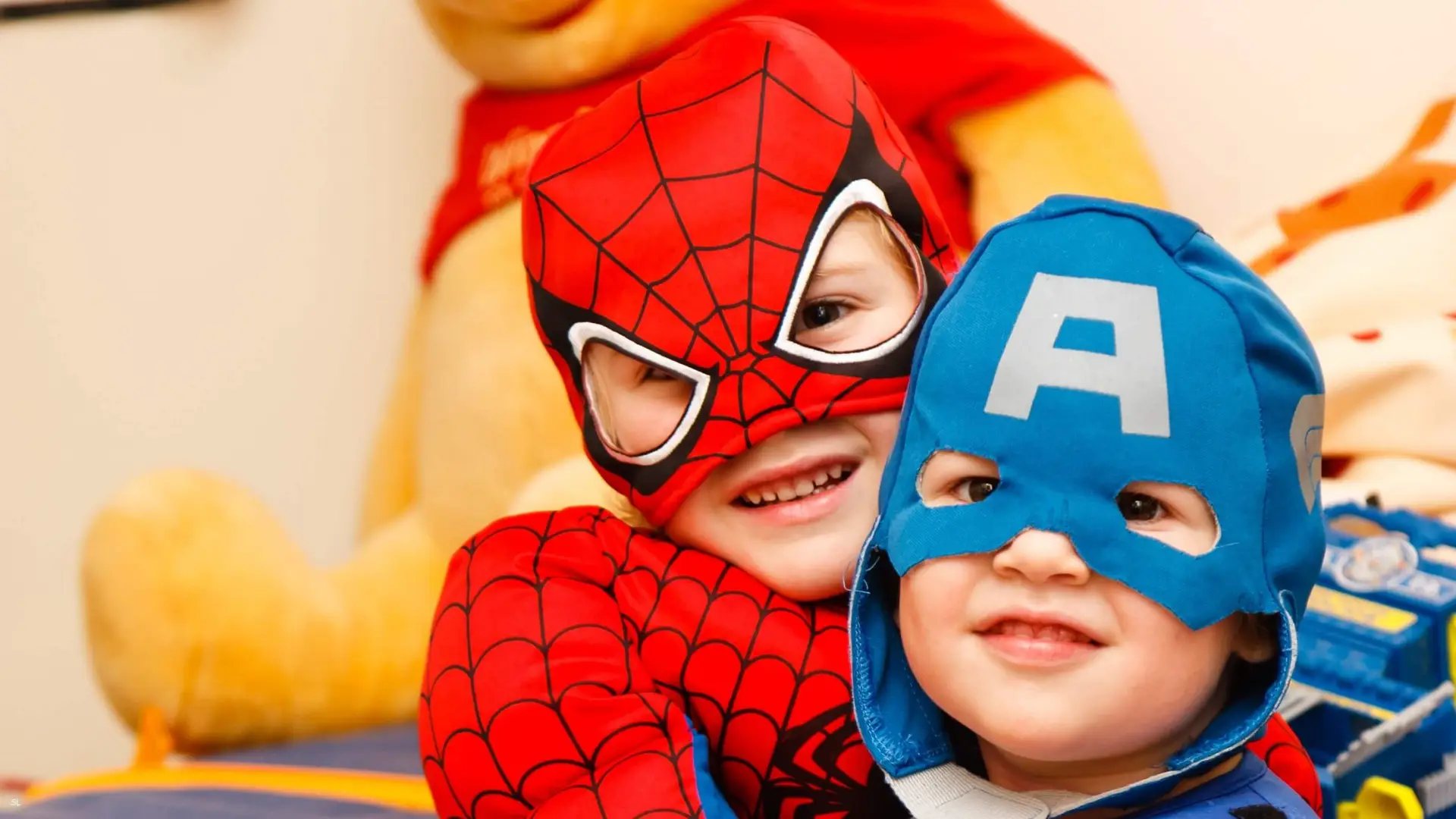 Two children in superhero costumes, one as Spider-Man and the other as Captain America, smile while sitting closely together, sharing a moment akin to stories found in books about making friends. A large stuffed animal is visible in the background.