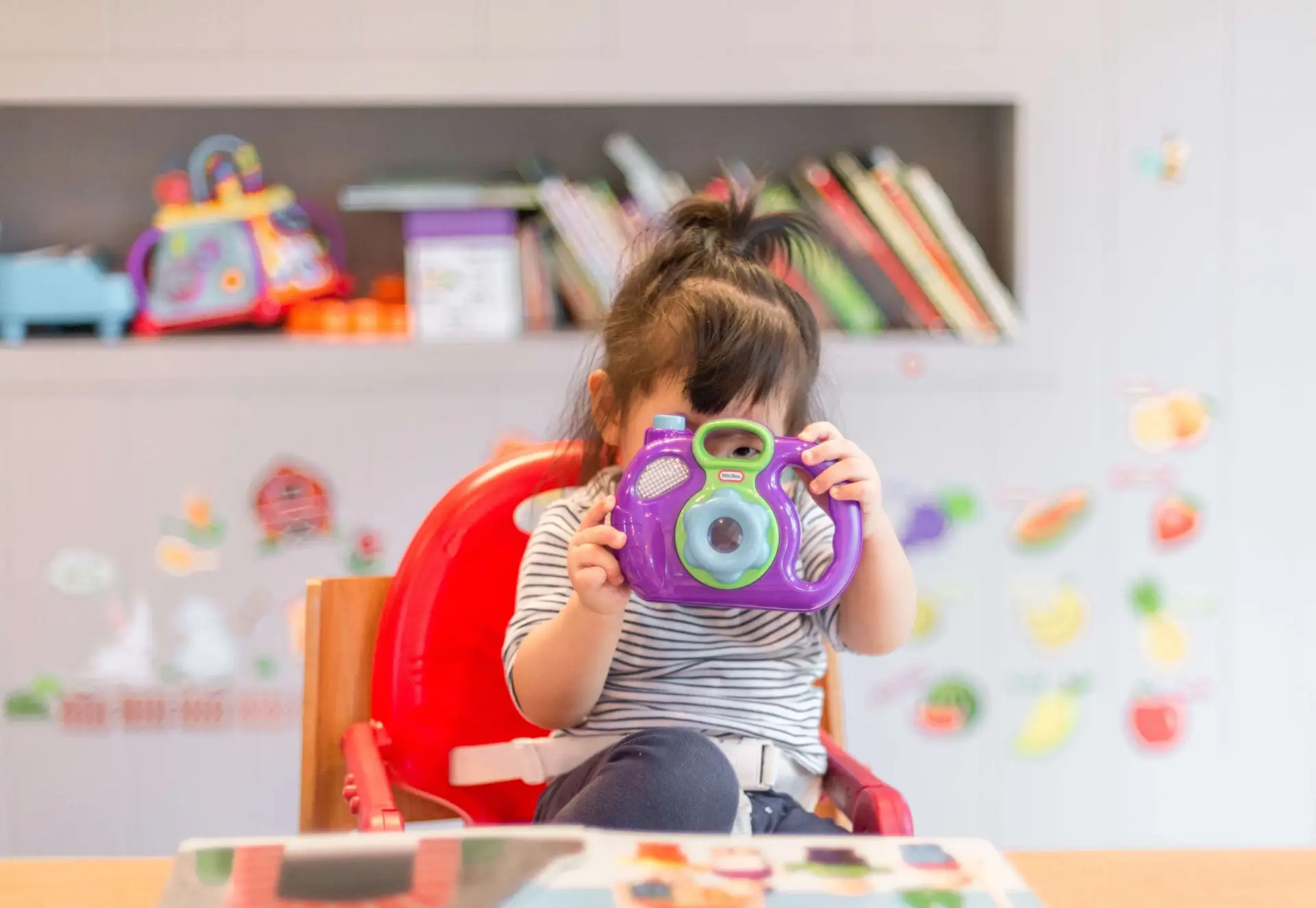 A young child with a ponytail sits in a red chair, holding a purple toy camera to their face. In the lively, child-friendly room, a shelf with books and colorful toys complements the vibrant kids activity table in the background.