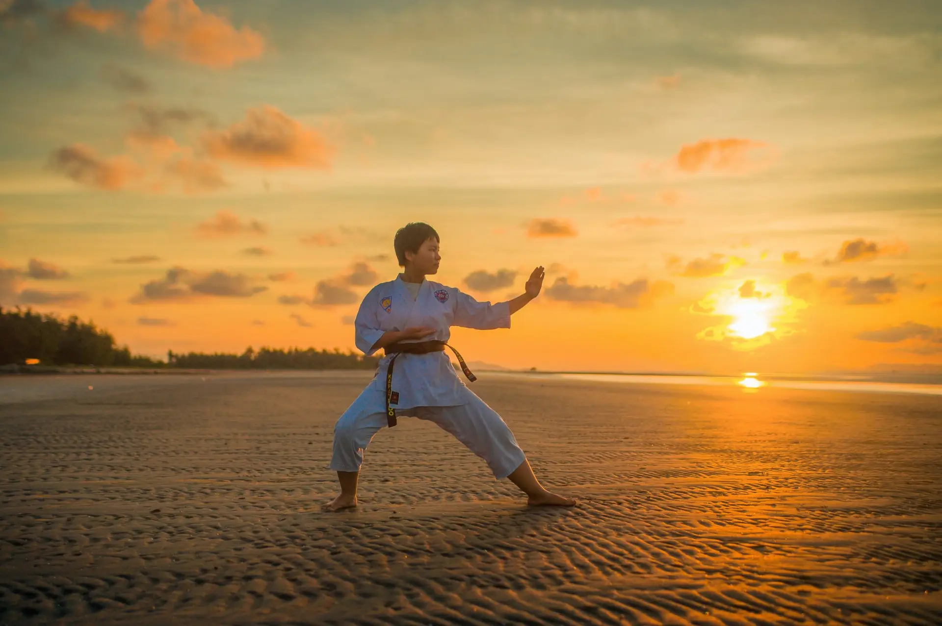 A person practices martial arts in a wide stance on a sandy beach at sunset, one of the best sports for kids with ADHD. The sky is filled with orange and pink hues, and the sun is near the horizon, casting a warm glow over the scene.