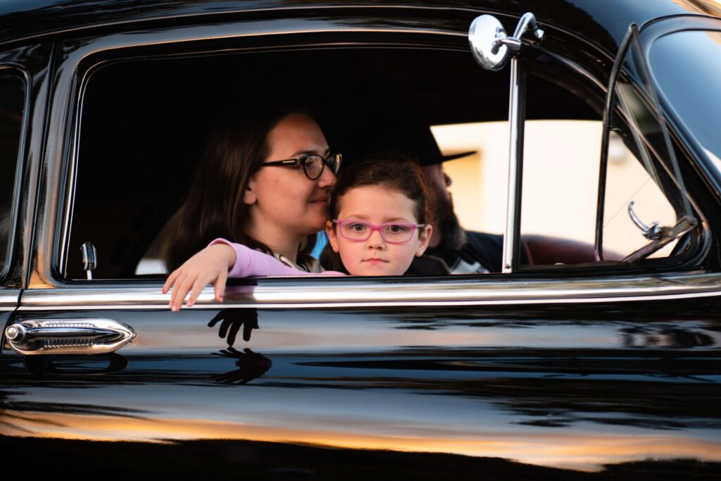 A woman and a young child wearing glasses sit inside a shiny black vintage car, with the child leaning out of the open window, perhaps imagining exciting car games for kids. Another person is partially visible behind them as the sunset reflects on the car's surface.