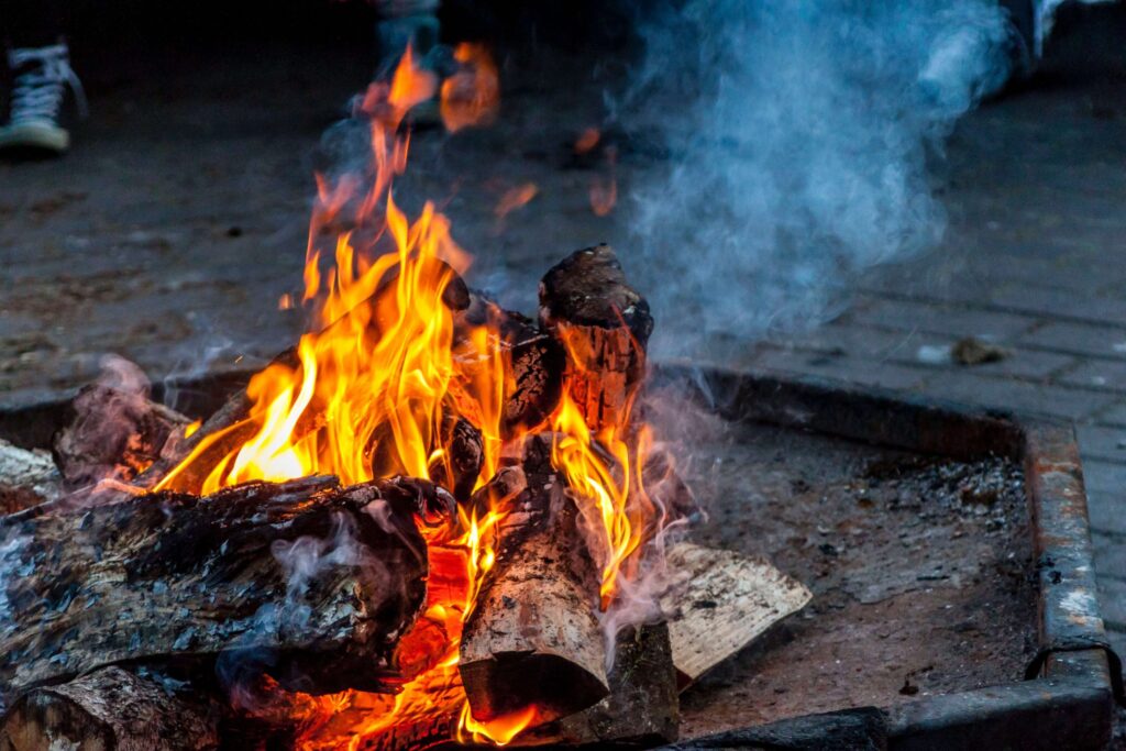 A close-up of a campfire with orange flames and smoke rising from burning logs, setting the perfect mood for campfire songs. The fire is contained within a square, metal fire pit on a stone surface.