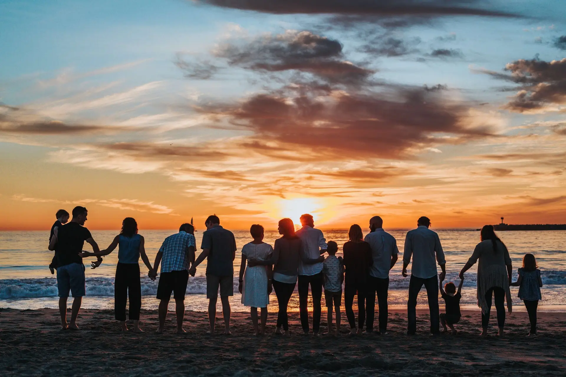 A group of people, including adults and children, stand holding hands on a beach, silhouetted against a colorful sunset sky. It's like a scene from books about family relationships, where ocean waves gently approach the shore, creating a serene and harmonious moment.