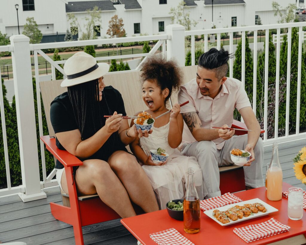 A family of three enjoys a meal on a patio. The child, sitting between a woman in a hat and a man, eagerly holds a bowl of kid-friendly chicken. The table has various dishes and drinks. They seem happy and relaxed in the outdoor setting.