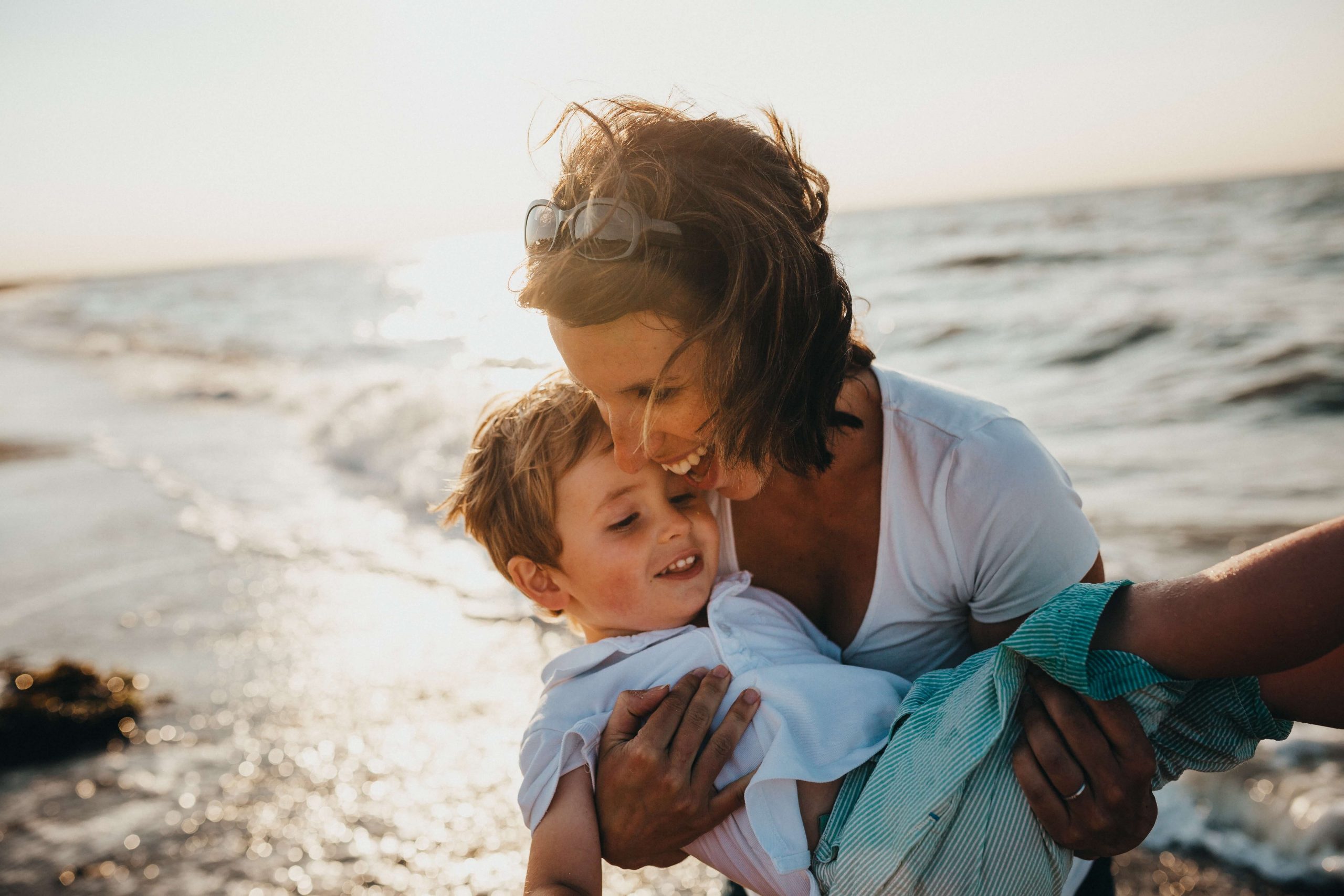 A woman joyfully holds a child as they both smile, standing on a sandy beach with gentle waves in the background. The sun is setting, casting a warm glow over the scene. Amid such peace, one might wonder why do kids bully when life offers such simple joys.