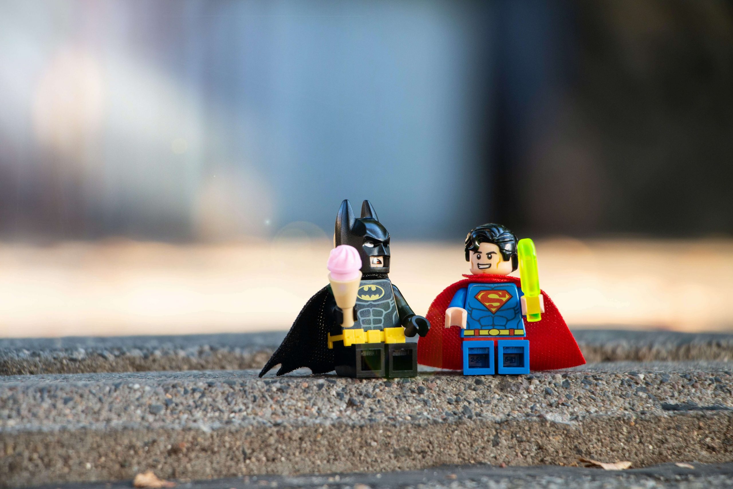 Two Lego figures, Batman and Superman, from beloved superhero movies for kids, sit together on a pavement. Batman holds an ice cream cone, while Superman grips a popsicle. The background is blurry, creating a casual and playful outdoor setting.