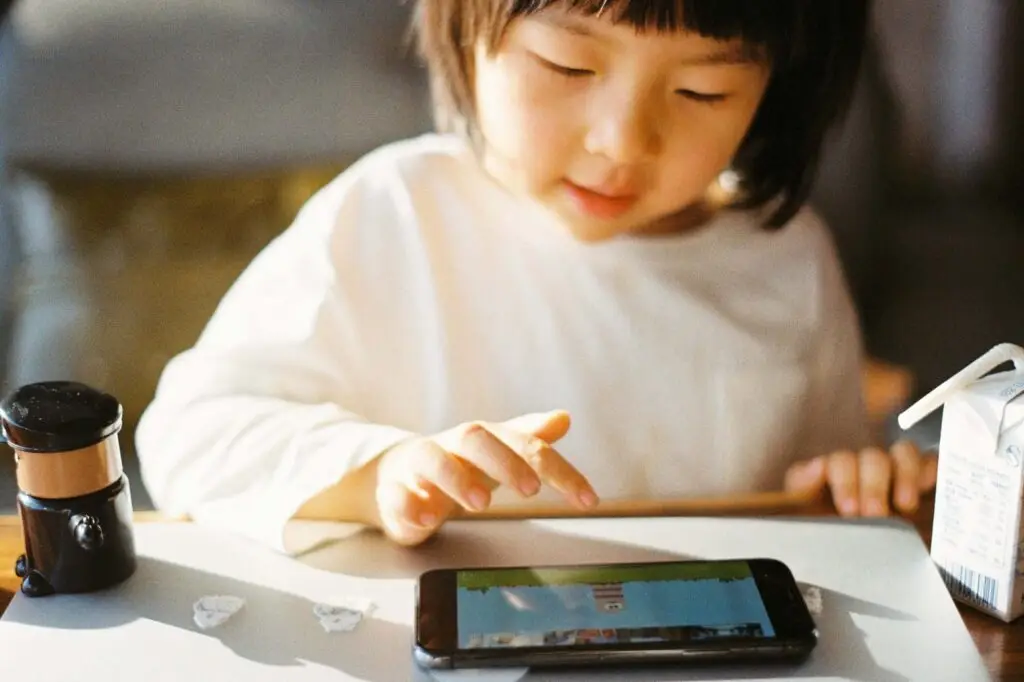 A young child in a white shirt plays an educational game on a smartphone, surrounded by playful props like a small toy and juice box. The cozy setting, enhanced by warm light, mirrors the vibrant charm of educational cartoons for kids.