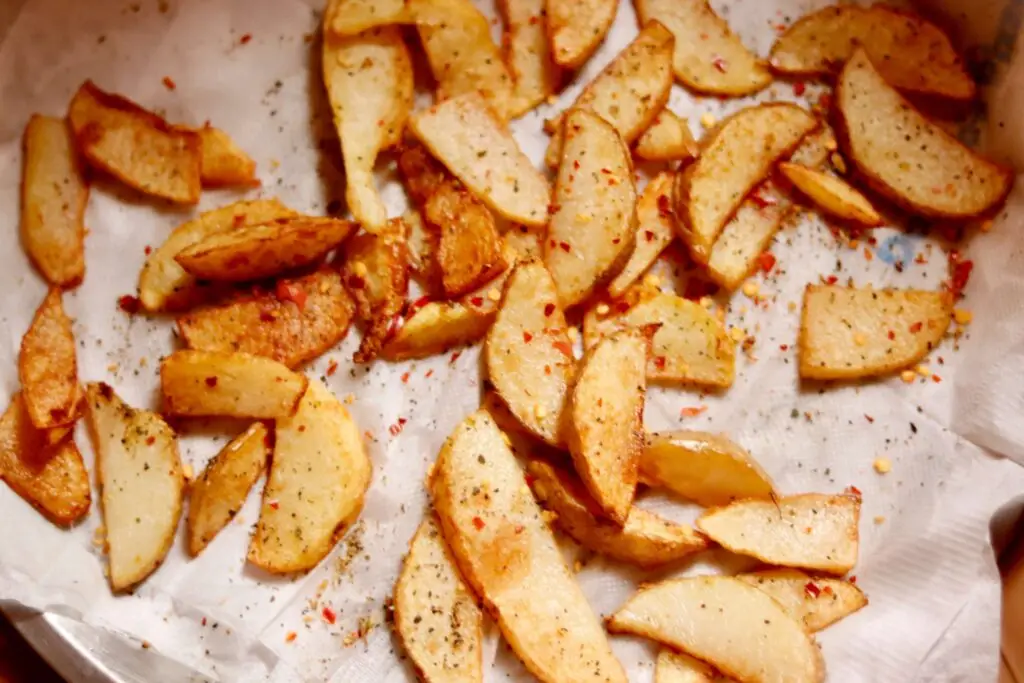 Close-up of seasoned potato wedges on parchment paper. These kid-friendly side dishes are golden brown, sprinkled with chili flakes and herbs, and arranged on a baking sheet, creating a crispy and appetizing appearance.