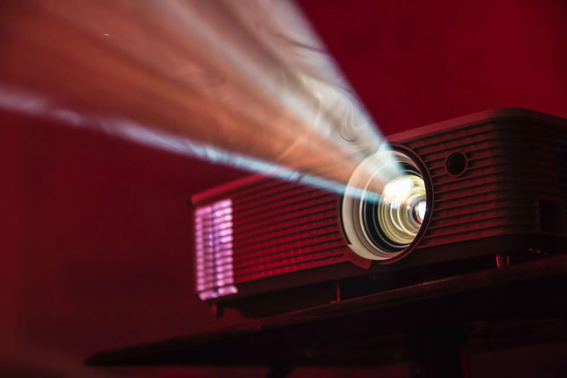 A projector emits a bright beam of light in a dark room with a red hue. The device, perfect for screening car movies for kids, is placed on a table, casting light forward, indicating its use for an engaging presentation or movie night.